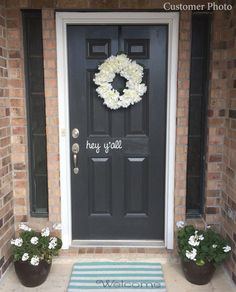 a black front door with white flowers and a welcome mat that says hey y'all