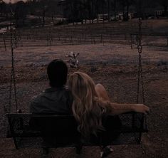 a man and woman sitting on a swing in the middle of a field at sunset