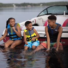 three children are sitting on the dock in front of a boat and one boy is wearing life vests