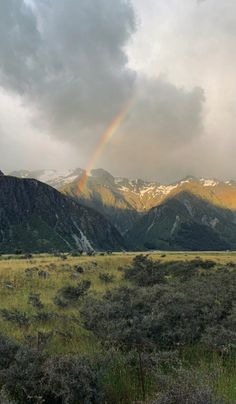 a rainbow shines in the sky over mountains and grass on a cloudy day,