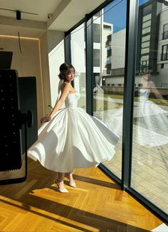 a woman in a white dress is standing near large windows