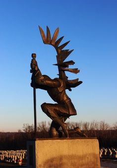 a statue of a man holding a spear on top of a cement block in the middle of a field