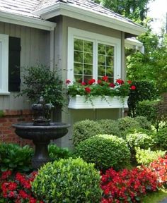 a garden with flowers, shrubs and a fountain in front of a house on a sunny day