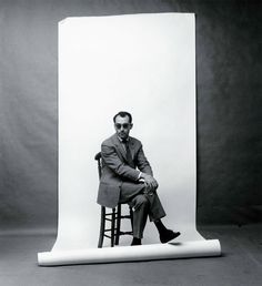 a black and white photo of a man sitting on a chair in front of a backdrop