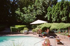 a woman sitting by the edge of a pool with an umbrella over her head,