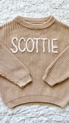 a knitted sweater with the word scottie written in white on it, sitting on a bed