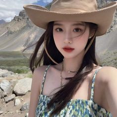 a young woman wearing a hat standing in front of some rocks and grass with mountains behind her