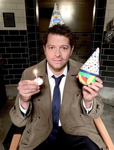 a man in a suit and tie holding two birthday hats on his fingers while sitting down