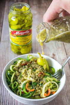 someone pouring olives into a bowl of pasta with pesto and peppers in it