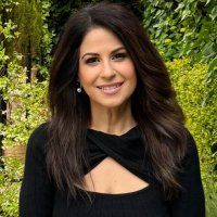 a woman with long dark hair wearing a black top and smiling at the camera, in front of greenery