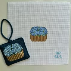 a cross - stitch card with a blue flowered cupcake on it and an embroidered ornament hanging from the front