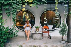 a woman sitting on a chair in front of two round windows with potted plants