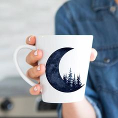 a woman holding a coffee mug with the moon and trees on it in front of her