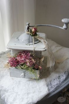 an old fashioned tea kettle with flowers in it sitting on a lace covered tablecloth