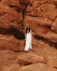 a woman is standing in front of some large rocks and wearing a long white dress