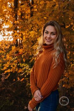 a woman in an orange sweater is posing for a photo near some trees with yellow leaves