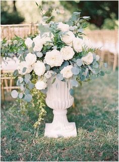 a white vase filled with flowers on top of a grass covered field next to chairs