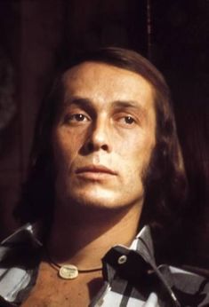 a man with long hair wearing a striped shirt and looking off to the side, in front of a dark background