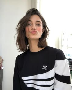 a woman with short brown hair wearing a black and white adidas sweater in front of a window