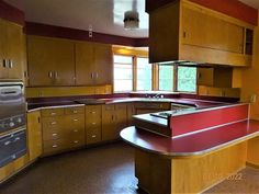 an empty kitchen with wooden cabinets and red counter tops on the island in front of two windows