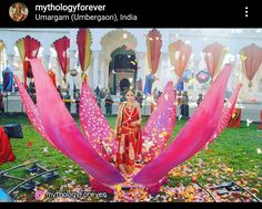 a woman in a red dress standing next to a large pink flower on top of a lush green field