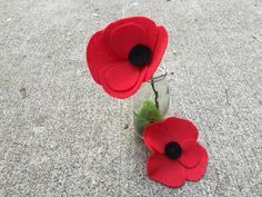 two red felt poppys in a jar on the ground next to some grass and dirt