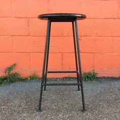 a black metal stool sitting in front of a brick wall with grass growing on the ground