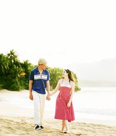 an older couple walking on the beach holding hands and smiling at each other, with palm trees in the background