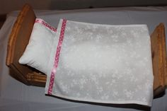 a wooden doll bed with white sheets and pink ribbon on the bottom half, sitting on a table