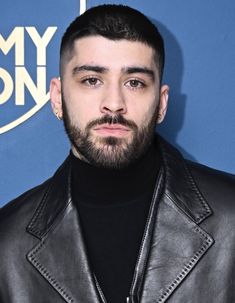 a man with a beard wearing a black turtle neck sweater and leather jacket standing in front of a blue wall