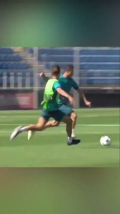 two men are running after a soccer ball on the field in front of an empty bleachers