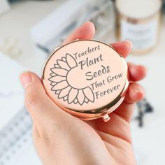 a person holding up a compact metal container with flowers on the front and words that read teachers plant seeds that grow forever