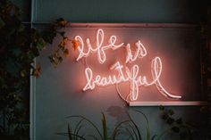 a neon sign that says life is beautiful on the side of a wall next to plants