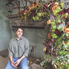 a man sitting on a stool in front of a fireplace with flowers hanging from it