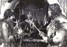 three men dressed in native american clothing standing next to each other and holding swords, talking