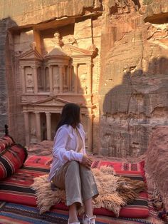 a woman sitting on top of a rug in front of a cliff side building with carved carvings