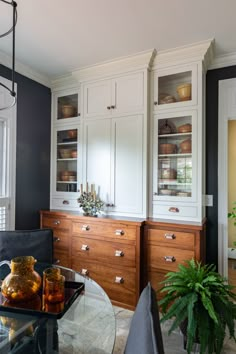 a dining room with glass table and white cupboards in the backround next to a potted plant