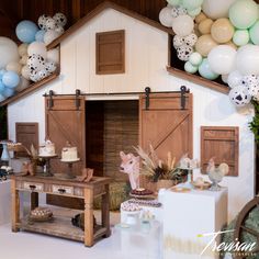 a table with cake and balloons in front of a barn - like backdrop for a baby's first birthday party