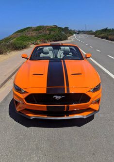 an orange sports car with black stripes driving down the road in front of a hill