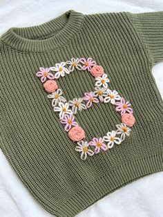 a green sweater with pink and white flowers on the front is laying on a bed