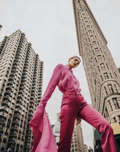 a woman standing in front of tall buildings wearing a pink suit and high heels with her hands on her hips