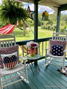 two white rocking chairs with red, white and blue pillows sit on a porch overlooking an american flag