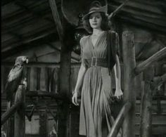 a woman in a dress and hat standing next to a parrot on a wooden platform