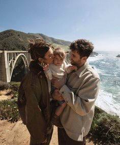 a man and woman holding a baby near the ocean with a bridge in the background