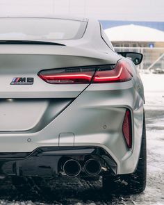 the rear end of a silver bmw car on snow covered ground with buildings in the background