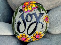 a rock with the word joy painted on it and flowers around it, sitting in front of some rocks