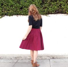 a woman standing on the sidewalk in front of a white wall wearing a maroon pleated skirt