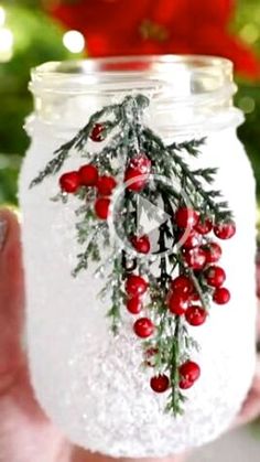 a hand holding a mason jar filled with snow and red berries on the inside, surrounded by christmas decorations