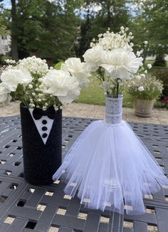 two vases with white flowers in them on a table outside, one has a tuxedo and the other is a bride's dress