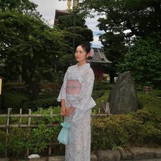 a woman in a white dress standing next to a light pole and bushes with trees behind her